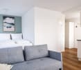 A small bedroom with a double bed, a gray sofa, a nightstand, a picture frame, and a kitchen area with a countertop and cabinets.