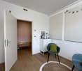 Empty room with wooden floor, white walls, open door, round rug, two chairs, white cabinet, and a small bed in the adjacent room.