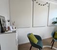 A room with a white wall, wooden floor, a white cabinet with a telephone, a vase, and a picture frame on top, two chairs with green pillows, and a round rug on the floor. There are three white boards mounted on the wall, and a string of green and white ba