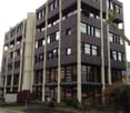 Multi-story office building with a modern, angular design featuring black and gray panels. The building has multiple floors and large windows. There is a small landscaped area in front of the building.