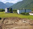 The image shows a residential area with multi-story apartment buildings surrounded by a grassy field and mountains in the background. There is an area of exposed soil and rocks in the foreground, suggesting some construction or landscaping work is underwa