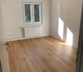 Empty room with white walls, wooden floor, and a window with a view of trees outside.