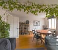 Living room with wooden floor, white walls, large windows, dining table, chairs, couch, bookshelves, plants, picture frame, dartboard, radiator