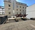 A multi-story building with multiple windows, a balcony, and a paved terrace with plants and a bench.