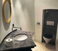 The image shows a bathroom with a granite vanity top, a round sink, a chrome faucet, a gold-framed mirror, and a black toilet. The floor is tiled, and there appears to be a dishwasher or similar appliance in the corner.