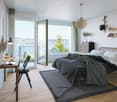 white walls, wooden floors, large bed, rug, nightstand with lamp, desk with book, chair, windows with balcony, view of ocean and trees