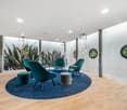 Wooden floor, white walls, blue round rug, green chairs, ottoman, round tables, green plant mural, glass windows