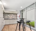 Modern kitchen with white cabinets, black countertops, stainless steel appliances, and a dining area with a high table and stools. Large windows provide a view of the outdoor greenery.