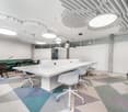 Modern and open office space with white desks, swivel chairs, and patterned carpet flooring. The ceiling features recessed lighting and a geometric design. The space also includes a glass partition wall and a view of the outdoors through a window.