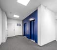 Empty hallway with white walls, gray carpet, two elevators, and two doors.