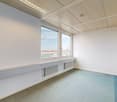 The image shows a spacious office space with large windows providing a view of the city skyline. The room has a clean, minimalist design with white walls, a suspended ceiling, and a tiled floor. There are radiators along the walls, suggesting this is a fu