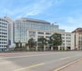 The image shows a modern glass and steel office building with a curved roof, surrounded by older, more traditional-style buildings. The building appears to be the Medizinischen Universität building, as indicated by the sign on the facade. The street in fr