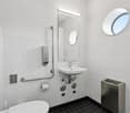 A toilet, sink, mirror, and a tissue dispenser, all with a modern and minimalist design. There is also a round window on the wall.