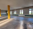 Open space with high ceiling, concrete floor, yellow columns, green strips on the walls, large windows with views to outside.
