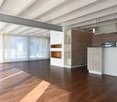 Open space with wooden floors, white walls, large windows, modern kitchen with white cabinets and countertops, wooden shelves, and a ceiling with exposed beams.