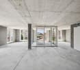 Large open space with concrete floors and ceiling, glass windows and doors, and minimal furnishings. The space appears to be a commercial or office area, ready for customization and renovation.