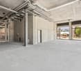 Large open commercial space with concrete floors, exposed ceiling, and brick walls. There is a metal security gate and a view of the exterior through the large windows.