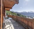 Outdoor balcony with wooden railings, two outdoor chairs, glass table, mountain views, wooden house.