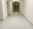 The image shows a long, narrow hallway with white walls and a concrete floor. The hallway has a series of recessed lighting fixtures in the ceiling, creating a well-lit and clean-looking space.