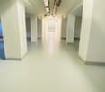 Long, narrow hallway with white walls and floors, recessed lighting, and pipes running along the ceiling.