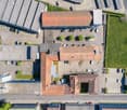 Aerial view of a large commercial building with a red tile roof, surrounded by a parking lot with several vehicles parked. The building appears to be a warehouse or industrial facility with multiple levels and loading docks. There is also some greenery, s