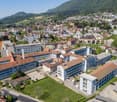 Aerial view of a city nestled in a valley surrounded by mountains. The city has a mix of modern and traditional buildings, with red-tiled roofs and multi-story structures. There are roads, parking lots, and green spaces visible. The landscape features for