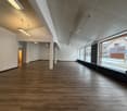 An empty room with hardwood floors, white walls, and fluorescent ceiling lights, large windows on one side. A signboard is positioned in front of the windows.
