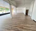 Large open space with hardwood-style flooring, white walls, and recessed lighting. The space appears to be a commercial or retail property with a large window overlooking the street.