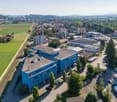Aerial view of a city with a mix of residential and commercial buildings, surrounded by green fields and hills. The city has a train station and various commercial and industrial buildings, including a large blue office building with a parking lot in the