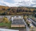 A large industrial building with green roofs, a spacious parking lot, surrounded by an extensive green field and forested hills in the background.