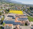 This image shows an aerial view of a town nestled in a valley surrounded by mountains. The town has a mix of residential and commercial buildings, with a large open field or agricultural area in the foreground. The buildings have a mix of architectural st