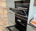 Integrated black oven with a glass door, open, in a kitchen with gray cabinets and a stone wall.