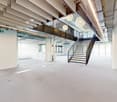 Large open-plan office space with high ceilings, exposed beams, and modern lighting fixtures. The space features a central staircase leading to a mezzanine level, and large windows providing natural light. The floors are covered in a light-colored concret