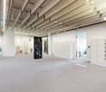 Large open office space with exposed ceiling beams, white walls, and lockers. There are various appliances and storage units visible, including what appears to be a dishwasher and a private washing machine.