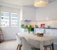 Dining room with white walls, wooden floor, white dining table, white chairs, white cabinets, large window, red shelf with plants and pitcher, flowers in vase, bowl of fruit, chandelier