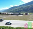 The image shows a large industrial or agricultural facility surrounded by fields and mountains. There are several large warehouse-like buildings and a few vehicles parked in the area. The landscape is dominated by green fields and hills in the background.