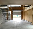 An empty garage with white tile walls, wooden beams, a large door, and a staircase.