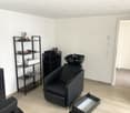 This image shows a room with a black salon chair, a black shelving unit, and a black cabinet. The room has white walls and a light-colored hardwood floor. There appears to be a private washing machine and a dishwasher visible in the room.