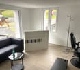 Spacious living room with large windows, hardwood flooring, black leather sofa, coffee table, and a white storage unit with shelves for displaying items.