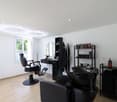 The image shows a hair salon with a modern and minimalist design. The room has white walls and a tiled floor. There is a large window that provides natural light and a view of the outdoors. The salon features black salon chairs, a styling station with a m