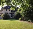 3-story house, yellow exterior, black roof, large balcony on the second floor, front garden with green grass and bushes