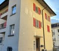 A multi-story residential building with a stucco exterior and red shutters on the windows. The building has a slanted roof and appears to be located in a mountainous area with a clear blue sky in the background.