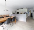 Dining room with wooden table, gray chairs, white walls, wooden floor, kitchen with white cabinets, microwave, refrigerator, and a statue