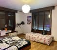 The image shows a bedroom with a large bed, a desk, and a white feather chandelier. The room has wooden furniture and windows, and the walls are decorated with artwork and posters.