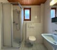 Bathroom with white tiles, a shower, toilet, and sink. The shower has a shower head and a soap dish. The toilet is white, and the sink is white with a faucet. There is a mirror on the wall above the sink.