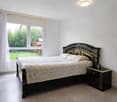 Bedroom with a double bed, nightstand, large window with white curtains, tiled floor, and a view of a grassy yard