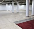 Large open commercial space with concrete flooring, white walls, and several white support columns. There is a red carpet in the center of the room.