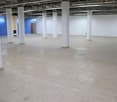The image shows a large, open commercial space with a concrete floor and white columns supporting the ceiling. The space appears to be unfinished or under renovation, with no visible furniture or decor. The walls are also white, and there is a blue door v