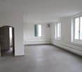 Large open-plan living space with white walls, gray tile flooring, and large windows providing natural light. The room appears to be empty, with no furniture or decor visible.