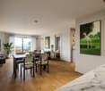 Dining room with wooden floor, large windows, dining table, chairs, couch, painting, potted plant, TV